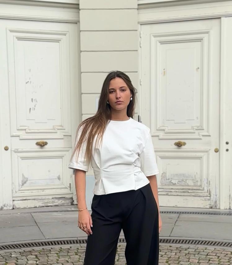 Femme en blouse blanche et pantalon noir Alibee Couture, posant devant une façade ancienne à Bruxelles.